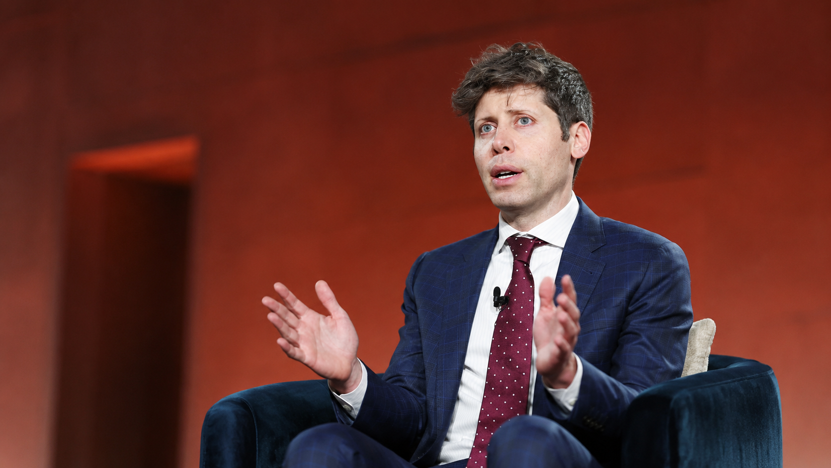 Sam Altman seduto su una poltrona durante un’intervista pubblica, con completo blu e cravatta bordeaux, mentre gesticola davanti a uno sfondo caldo arancione