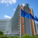 Sede della Commissione Europea a Bruxelles con bandiere dell’Unione Europea in primo piano, cielo azzurro e architettura istituzionale moderna.