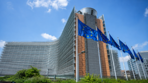 Sede della Commissione Europea a Bruxelles con bandiere dell’Unione Europea in primo piano, cielo azzurro e architettura istituzionale moderna.