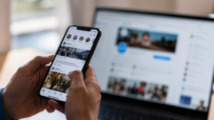 Mani che scorrono il feed di un social network su uno smartphone, con lo schermo di un computer sfocato sullo sfondo e luce naturale morbida.