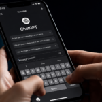 Primo piano di uno smartphone con l’app ChatGPT aperta in modalità scura, mentre una persona digita sulla tastiera. Sfondo scuro e illuminazione da studio, in stile editoriale tecnologico.