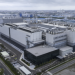 Vista aerea di un grande impianto industriale giapponese per la produzione di memorie flash, con ampi edifici produttivi, sistemi di ventilazione e infrastrutture tecnologiche sotto un cielo grigio.