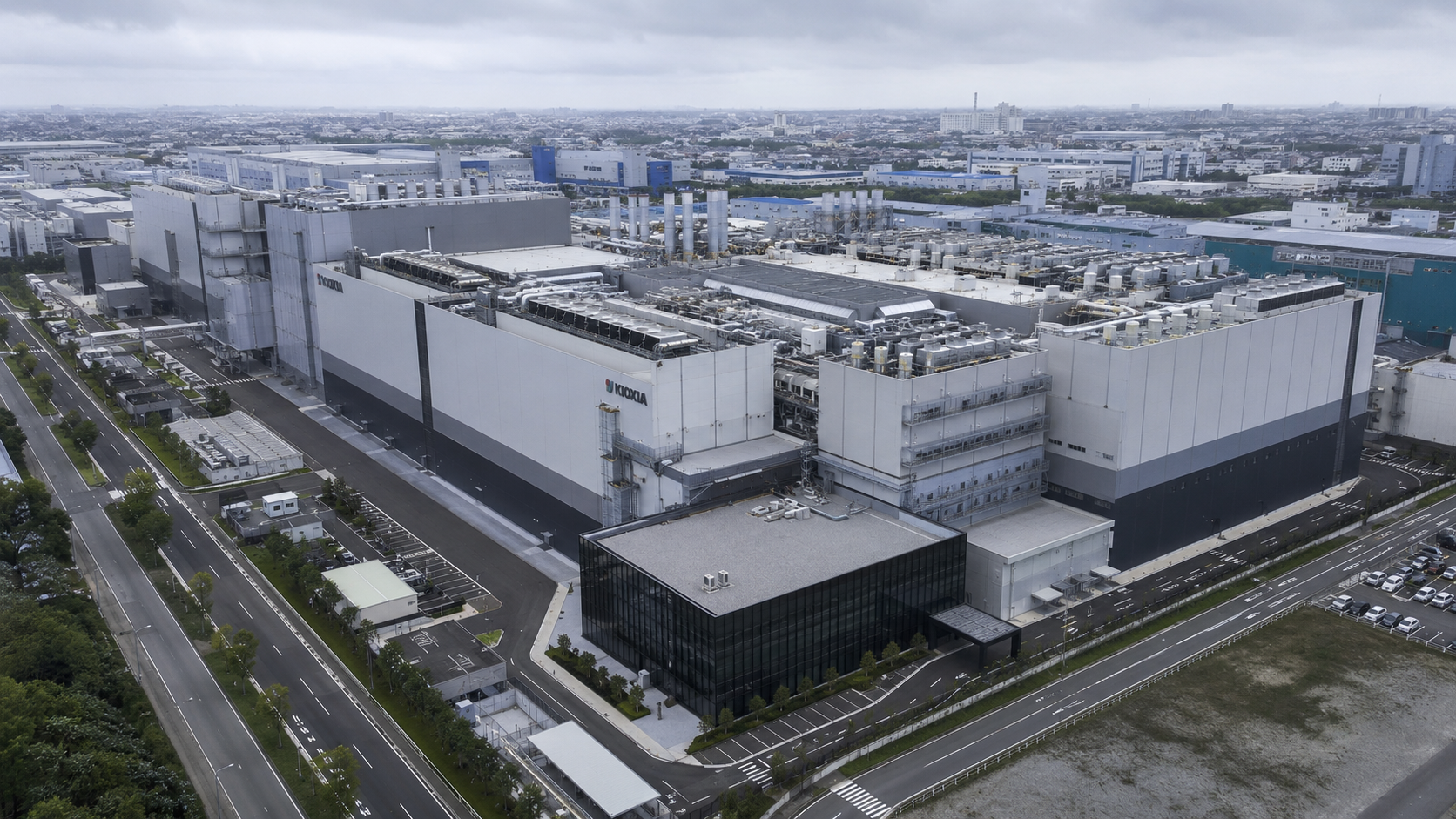 Vista aerea di un grande impianto industriale giapponese per la produzione di memorie flash, con ampi edifici produttivi, sistemi di ventilazione e infrastrutture tecnologiche sotto un cielo grigio.