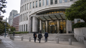 Esterno di un moderno tribunale federale a Oakland, con alcune persone in abiti formali dirette verso l’ingresso.