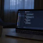 Laptop aperto su una scrivania in penombra con schermata di chat, tastiera con caratteri cirillici e luce fredda azzurra in una stanza con tende chiuse.