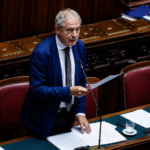 Ministro Adolfo Urso durante un intervento alla Camera dei Deputati, in piedi tra i banchi dell’aula parlamentare con un documento in mano.