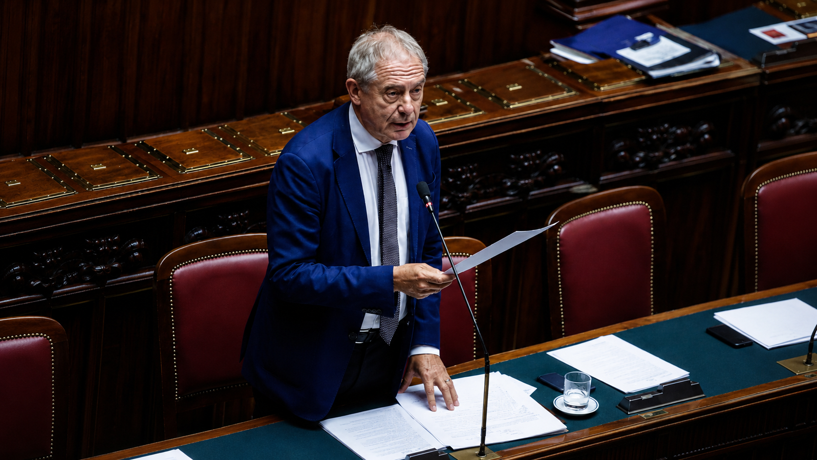 Ministro Adolfo Urso durante un intervento alla Camera dei Deputati, in piedi tra i banchi dell’aula parlamentare con un documento in mano.