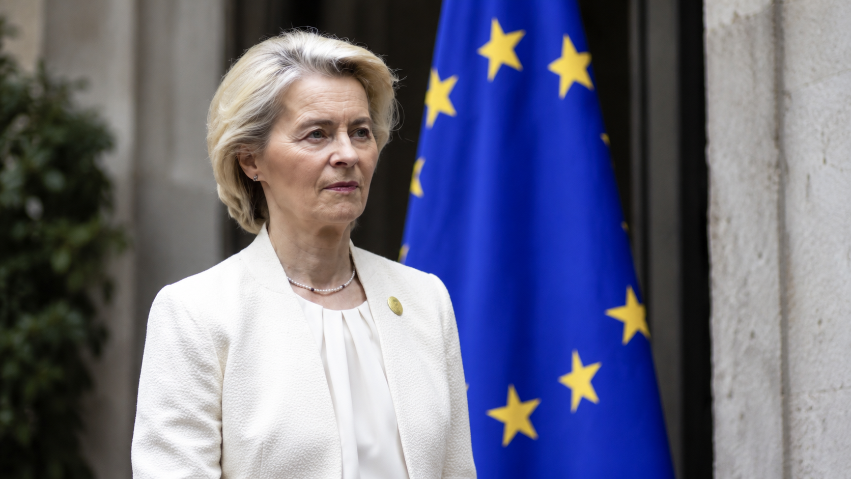 Donna in abito chiaro fotografata all’aperto accanto a una bandiera dell’Unione europea, in un ritratto istituzionale realistico con sfondo architettonico sfocato.