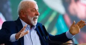 Il presidente brasiliano Luiz Inácio Lula da Silva interviene durante un discorso pubblico, con le braccia aperte davanti ai microfoni. Sullo sfondo, sfocati, i colori della bandiera brasiliana.