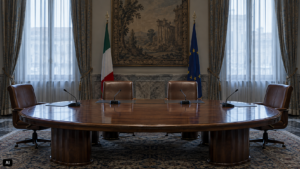 Sala riunioni istituzionale vuota con tavolo ovale in legno, quattro poltrone in pelle e microfoni spenti. Luce fredda dalle finestre, bandiere italiana ed europea sullo sfondo