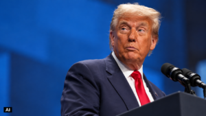 Donald Trump ripreso di tre quarti davanti ai microfoni, con abito blu e cravatta rossa, su sfondo blu. Immagine rielaborata con IA, con etichetta “AI” in basso a sinistra