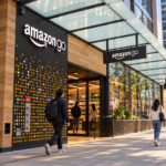 Ingresso di un negozio Amazon Go visto da una prospettiva laterale, con insegne del marchio, vetrate, passanti e una strada urbana sullo sfondo.