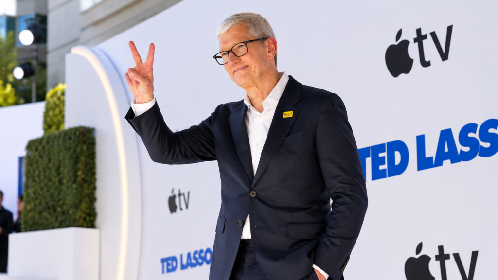 Tim Cook sul red carpet di Ted Lasso con completo scuro, occhiali e gesto della vittoria davanti al backdrop Apple TV.
