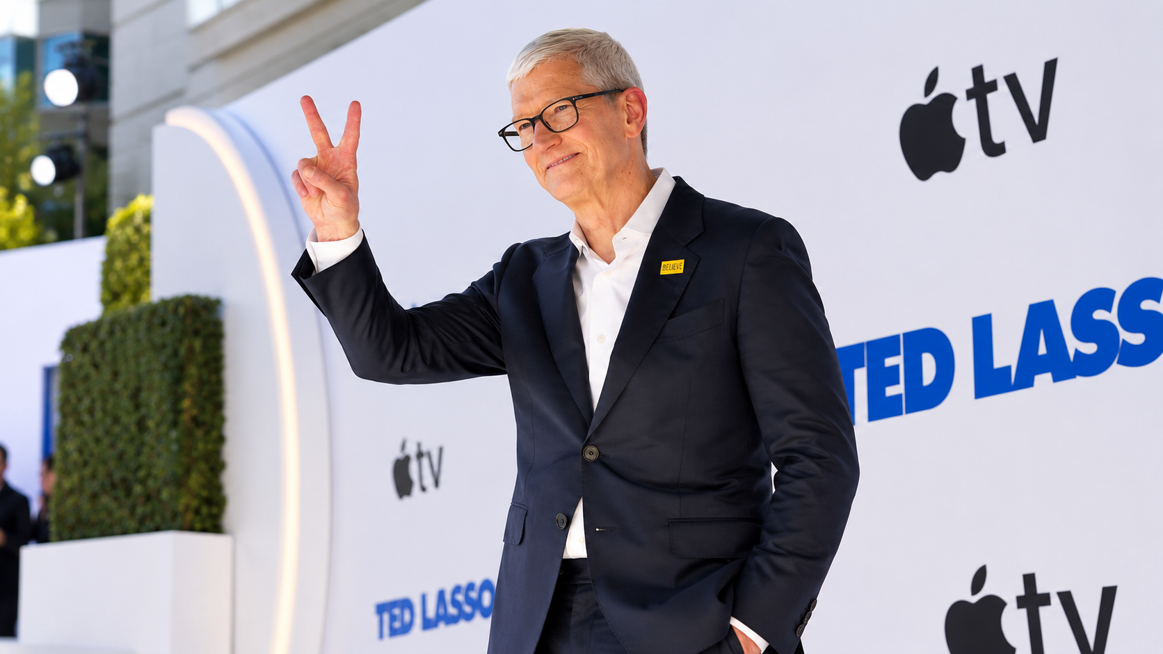 Tim Cook sul red carpet di Ted Lasso con completo scuro, occhiali e gesto della vittoria davanti al backdrop Apple TV.